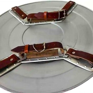 May include: A circular, silver-coloured shield featuring brown leather straps and metal brackets. The shield has two sets of straps for arm security. The leather straps are riveted to the metal brackets.