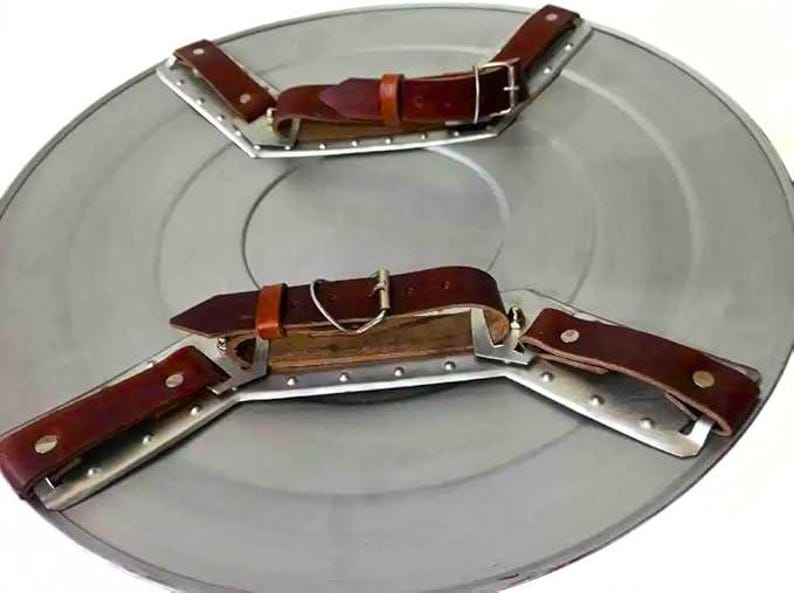 May include: A circular, silver-coloured shield featuring brown leather straps and metal brackets. The shield has two sets of straps for arm security. The leather straps are riveted to the metal brackets.