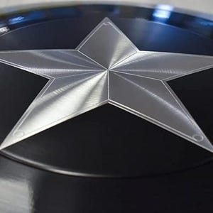 May include: A close-up of a black and silver shield. The shield features a large, five-pointed silver star in the centre against a black background. The star has a textured appearance, and the shield has a glossy finish.