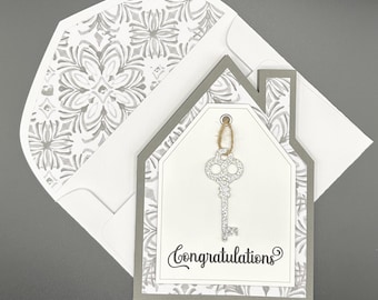 May include: A handmade greeting card with a house-shaped design. The card features a silver glitter key charm hanging from a gold string. The card reads "Congratulations" in elegant script. The envelope has a white and gray floral pattern.