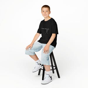 Op de afbeelding: Een zwart t-shirt met de tekst "No hockey today" en een stokfiguur met een verdrietig gezicht. De persoon draagt lichtblauwe spijkerbroekshorts en witte sneakers. De persoon zit op een zwarte kruk.