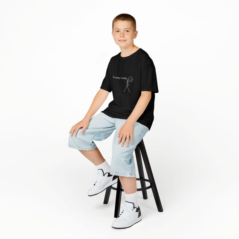 Op de afbeelding: Een zwart t-shirt met de tekst "No hockey today" en een stokfiguur met een verdrietig gezicht. De persoon draagt lichtblauwe spijkerbroekshorts en witte sneakers. De persoon zit op een zwarte kruk.