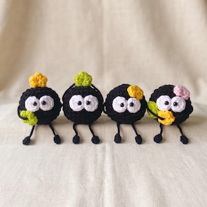 May include: Four round, black crocheted figures with dangling legs and large white eyes. Each figure features a small, colorful crocheted accent on top or side, including yellow, lime green, and pink floral or star-shaped embellishments.