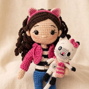 May include: A handcrafted crochet doll with dark brown curly hair, wearing a pink headband with cat ears, a bright pink jacket, a black and white striped shirt, and blue pants. The doll holds a small white and gray crochet cat toy with a pink bow.