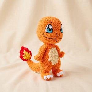 Puede incluir: Un peluche tejido a ganchillo de un personaje de lagarto bípedo hecho con hilo naranja brillante, con una mancha de color crema en el vientre y grandes ojos azules. La criatura tiene una cola larga que termina en un detalle de hilo rojo y amarillo.
