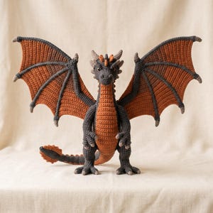 May include: A crocheted dragon figurine featuring a dark charcoal gray body with a burnt orange underbelly and wing membranes. The creature has large, spread wings, a long tail with a spiked tip, and small horns on its head, all crafted in yarn.