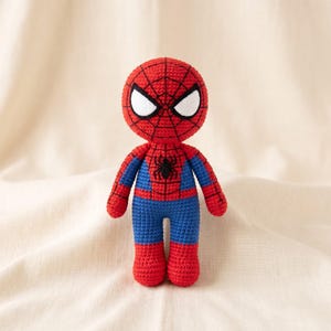 Könnte beinhalten: Eine handgefertigte Häkelpuppe einer Superheldenfigur in den Farben Rot und Blau. Der Kopf hat ein schwarzes Spinnennetzmuster auf rotem Garn mit großen weißen Augen. Ein kleines schwarzes Spinnenemblem befindet sich mittig auf dem blauen Oberkörper.