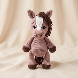 May include: A crocheted horse stuffed toy in a dusty rose color with a white blaze on its face. It features dark brown yarn for the mane, tail, and hooves, along with two black safety eyes. The toy has a rounded body, pointed ears, and limbs.