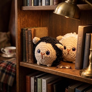 May include: Two round, plush, crocheted sheep figures sit on a wooden bookshelf. One sheep has black wool and a cream face, while the other is entirely cream-colored. Both have large, dark, glossy eyes and small, stitched noses, positioned near books.