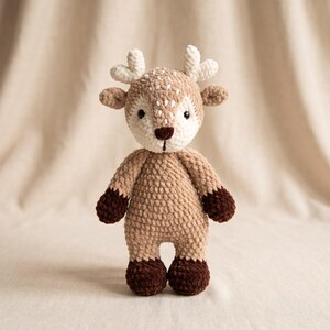 May include: A crochet deer plush toy featuring a light tan body, a cream-colored face with white spots, and dark brown hooves. The toy has small ears, white antlers, and a dark brown nose. The texture is soft and fuzzy, with visible yarn stitches.