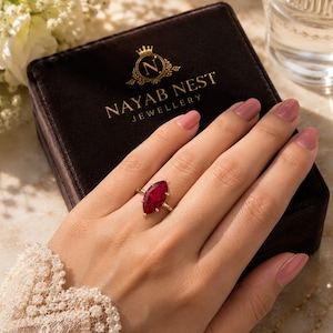 May include: A gold ring featuring a marquise-cut deep red gemstone, worn on a finger. The ring is positioned over a dark brown velvet jewellery box with the gold text NAYAB NEST JEWELLERY and a crown logo printed on the lid surface.