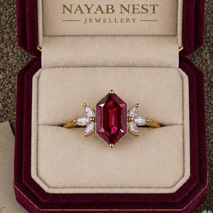 May include: A gold ring featuring a large, hexagonal, deep pink gemstone flanked by two clusters of three marquise-cut white stones. The ring is displayed in a dark red velvet box with the text NAYAB NEST JEWELLERY printed on the beige interior.