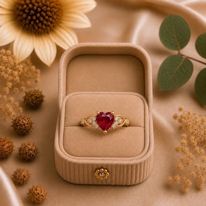 Puede incluir: Un anillo de oro con una piedra preciosa roja en forma de corazón en el centro, flanqueada por pequeñas piedras claras a cada lado. El anillo se presenta dentro de un joyero de terciopelo beige de forma rectangular redondeada con un cierre.