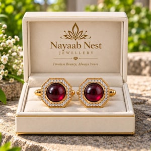 Puede incluir: Un par de gemelos octogonales dorados con una piedra cabujón de color rojo intenso en el centro rodeada de pequeños cristales transparentes. Los gemelos se presentan en un joyero color crema con el texto Nayaab Nest Jewellery impreso.