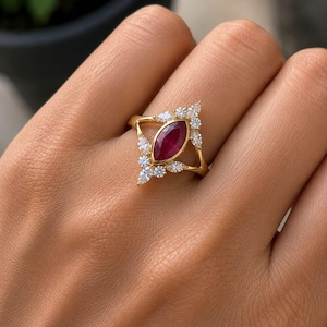 Op de afbeelding: Een gouden ring met een centrale dieprode robijn in marquise-slijpvorm. De robijn is omringd door een cluster van kleine, heldere, peervormige en rondgeslepen witte stenen in een symmetrisch, puntig ontwerp langs de ringband.