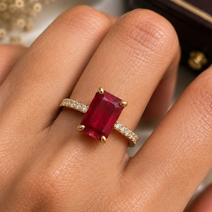 May include: A gold ring featuring a large, rectangular, deep red gemstone set in four prongs. The band is crafted from yellow gold and is adorned with a series of small, round, clear white diamonds set along the sides of the ring shank.