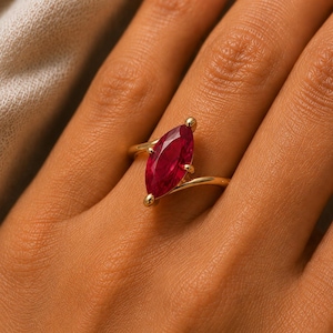 Könnte beinhalten: Ein Goldring mit einem einzelnen tiefroten Edelstein im Marquise-Schliff, der in einer Ringschiene im Bypass-Stil gefasst ist. Der leuchtend rote Stein ist vertikal ausgerichtet, wird von kleinen Krappen gehalten und das polierte Goldband.
