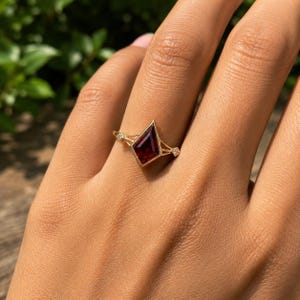 Peut inclure: Une bague en or avec une pierre précieuse rouge foncé en forme de cerf-volant montée en serti clos. L'anneau est fin et divisé, orné de deux petites pierres claires de taille carrée de chaque côté de la pierre centrale rouge grenat.