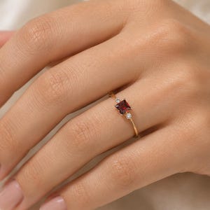 Puede incluir: Un anillo de oro fino con una piedra preciosa de granate rojo oscuro de corte princesa en el centro, flanqueada por dos pequeñas piedras de acento redondas y transparentes. La banda es delgada y pulida, con un diseño delicado y engaste.
