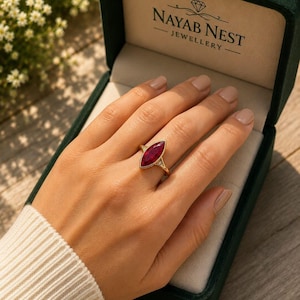 Puede incluir: Un anillo de oro con una piedra preciosa de color rojo intenso en talla marquesa montada en una banda con pequeñas piedras de acento. El anillo se presenta en un joyero de terciopelo verde oscuro con el texto NAYAB NEST JEWELLERY y un icono de diamante.
