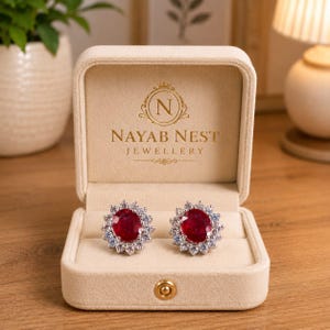 Puede incluir: Un par de pendientes de botón con una piedra roja de corte ovalado rodeada por un halo de pequeñas piedras transparentes, presentados en un joyero de terciopelo beige. La tapa interior muestra el texto dorado NAYAB NEST JEWELLERY.