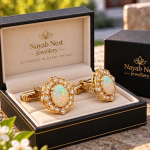 Op de afbeelding: Een paar gouden manchetknopen met ovale opalen omringd door kleine heldere stenen in een zeshoekige zetting. De manchetknopen worden gepresenteerd in een zwart sieradendoosje met de tekst Nayab Nest Jewellery in goud op het deksel.