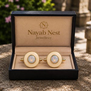May include: A pair of round gold-tone cufflinks featuring a central iridescent opal-like stone surrounded by a smooth cream-colored border. The cufflinks are presented in a black hinged box with the text Nayab Nest Jewellery printed inside.