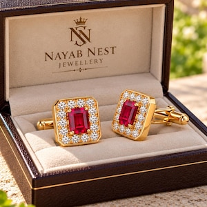 May include: A pair of gold cufflinks featuring rectangular red gemstones surrounded by small white stones, presented in a dark brown box with the text NAYAB NEST JEWELLERY printed in gold on the interior lid of the open jewelry case.