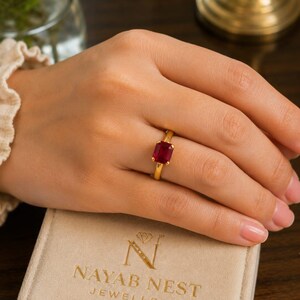 Può includere: Un anello in oro con una pietra preziosa rosso intenso dal taglio rettangolare a smeraldo, fissata con quattro griffe. L'anello poggia su un portagioie beige con la scritta NAYAB NEST JEWELLERY e un'icona a forma di diamante sopra la N.