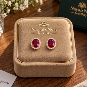 May include: A pair of oval-shaped stud earrings featuring deep red center stones surrounded by a halo of small white crystals. The earrings are displayed on a tan velvet jewelry box with the gold text Nayab Nest Jewellery printed on top.