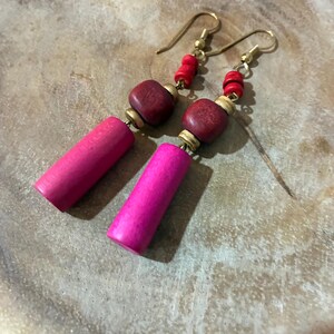 Puede incluir: Pendientes hechos a mano con una combinación de cuentas de madera rosa vibrante y rojo intenso. Los pendientes tienen ganchos dorados y pequeñas cuentas rojas. El diseño incluye cuentas cilíndricas y cuadradas, creando un accesorio único y llamativo.