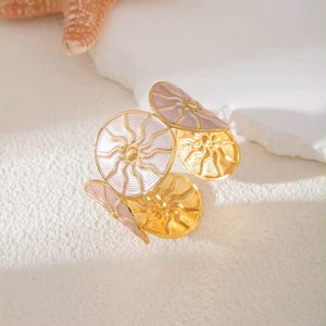 May include: A gold-toned metal ring featuring three circular motifs with a sunburst pattern. Each circle has a pale pink enamel inlay with raised gold lines radiating from the center. The band is open, allowing for adjustment, and has a polished finish.
