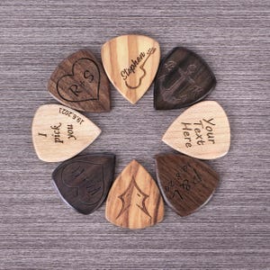 Könnte beinhalten: Eine Sammlung personalisierter Gitarren-Plektren aus Holz, kreisförmig angeordnet. Die Plektren weisen Gravuren auf, darunter Initialen, Daten und die Texte "Stephen", "I pick you" und "Your Text Here". Die Plektren sind aus verschiedenen Holzarten gefertigt.
