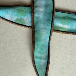 Może przedstawiać: Dwie wstążki z wzorem tie-dye w odcieniach niebieskiego i zielonego. Krawędzie wykończone brązową obw&oacute;dką i ciemnoniebieskim ściegiem.