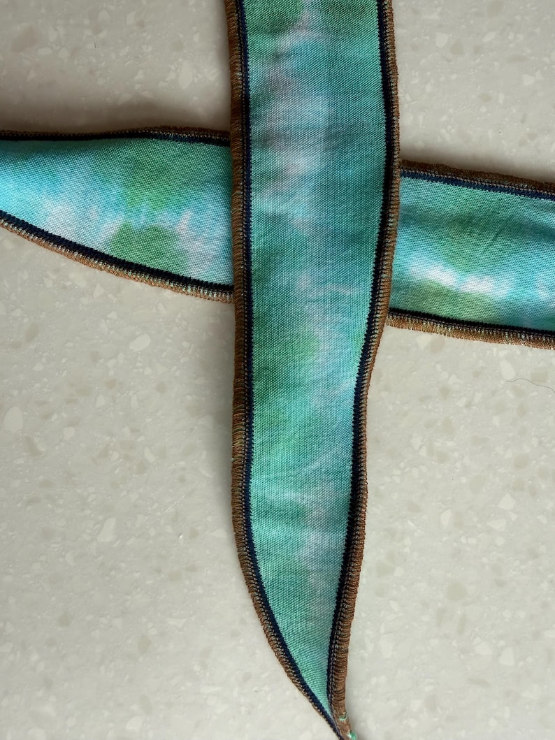 Może przedstawiać: Dwie wstążki z wzorem tie-dye w odcieniach niebieskiego i zielonego. Krawędzie wykończone brązową obw&oacute;dką i ciemnoniebieskim ściegiem.