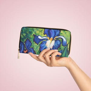 May include: A rectangular zip-around wallet featuring a vibrant, painterly print of deep blue iris flowers with white and yellow centers set against a lush green foliage background. The wallet has a gold-toned metal zipper pull and black trim.