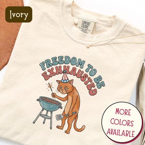 Puede incluir: Camiseta de manga larga color marfil con un gráfico de un gato con un gorro de fiesta, sosteniendo una bengala y una espátula cerca de una parrilla. El texto dice "Freedom to be Exhhausted". Una pequeña etiqueta dice "Comfort Colors".