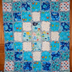 May include: A blue and white patchwork quilt with a nautical theme. The quilt is made of various fabrics with different patterns, including whales, fish, and seashells. The quilt is edged with a white fringe.