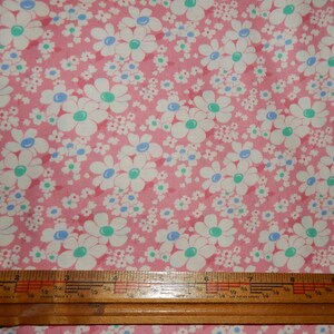 May include: Pink fabric with a repeating floral pattern. Large white daisies with teal and blue centers are scattered across the fabric, along with smaller white flowers. A wooden ruler is at the bottom of the image.