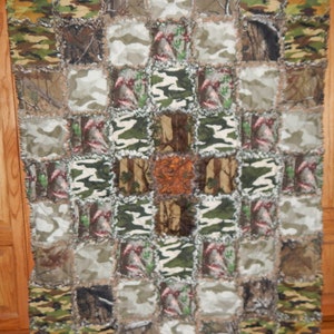 May include: A patchwork quilt made of various camouflage fabrics in shades of green, brown, and white. The quilt is made up of squares and is sewn together with a frayed edge.