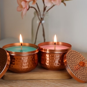 May include: Two lit candles in copper-colored containers with lids. One candle has teal wax, the other pink. The lids are off to the side. The candles sit on a wooden surface with flowers in the background.
