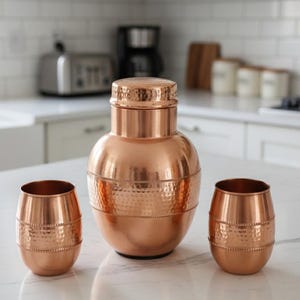 Könnte beinhalten: Ein kupferfarbenes Bar-Set mit einem großen Shaker mit Deckel und zwei passenden Bechern. Der Shaker hat ein strukturiertes, gehämmertes Design in der Mitte. Das Set wird auf einer weißen Arbeitsplatte in einer Küche präsentiert.