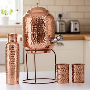 May include: A copper water dispenser with a hammered texture, a matching water bottle, and two small cups. The dispenser sits on a brown metal stand. The set is displayed on a white surface in a kitchen setting.