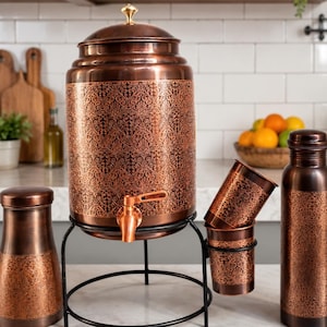 May include: A copper-colored beverage dispenser with a floral pattern, a spigot, and a lid with a gold-colored finial. It is accompanied by matching cups, a bottle, and a smaller container. The dispenser sits on a black metal stand.