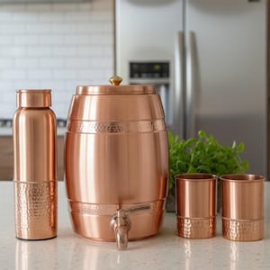 May include: A copper water dispenser set, including a barrel-shaped dispenser, a water bottle, and two tumblers. The dispenser has a tap and a hammered texture on the base. The set is made of copper and is displayed on a countertop.