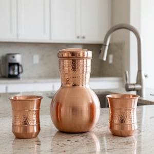 May include: A copper water jug with a hammered texture, a matching lid, and two copper cups. The jug has a bulbous body and a cylindrical top. The cups have a hammered texture and a flared rim. The set is displayed on a countertop.