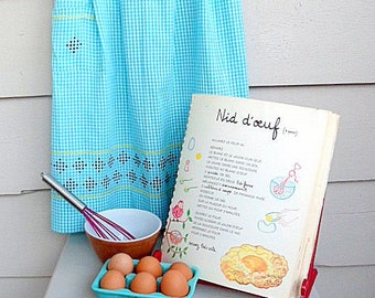 Op de afbeelding: Een vintage keuken scène met een blauw geruite schort, een kookboek getiteld "Nid d'oeuf", een kom, een garde en een eierdoos. De schort heeft decoratieve randen. Het kookboek is open voor een recept.