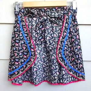 May include: A black floral apron with a pink and blue trim. The apron has a bow at the waist and a scalloped edge.