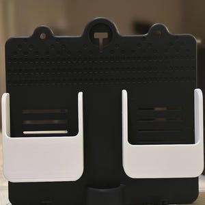 May include: A black and white wall organiser with two white storage compartments and a small bottom holder. The organiser has a perforated surface and a 'T' shaped design element at the top.