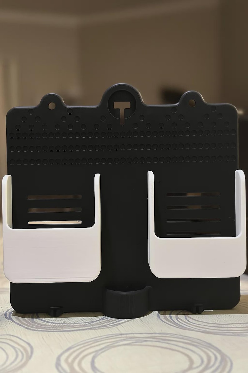 May include: A black and white wall organiser with two white storage compartments and a small bottom holder. The organiser has a perforated surface and a 'T' shaped design element at the top.